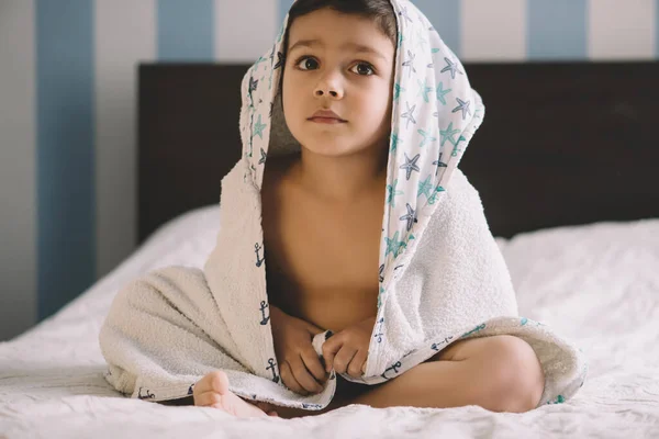 Милый ребенок, покрытый полотенцем с капюшоном, сидит на кровати и смотрит в сторону — стоковое фото