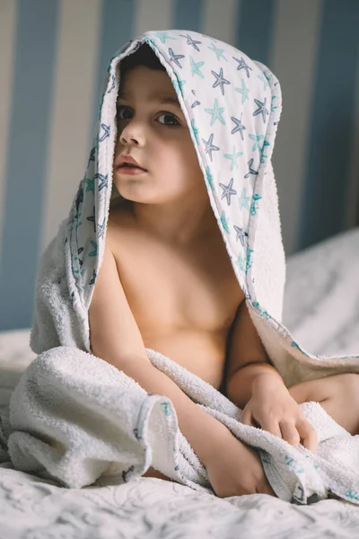 Lindo niño, envuelto en una toalla con capucha, sentado en la cama y mirando a la cámara - foto de stock