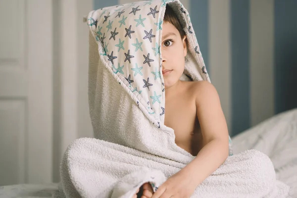 Adorable garçon, couvert de serviette à capuche, assis sur le lit et regardant la caméra — Photo de stock