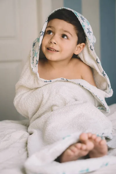Selective focus of happy child, wrapped in hooded towel, looking away while sitting on bed — Stock Photo