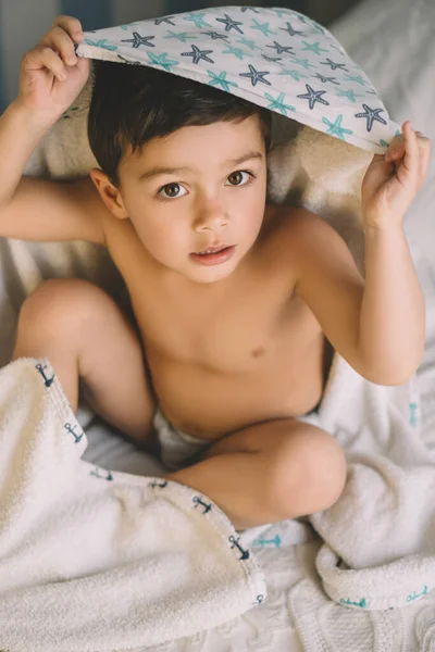 Vista aérea del niño lindo, envuelto en una toalla con capucha, sentado en la cama y mirando a la cámara - foto de stock