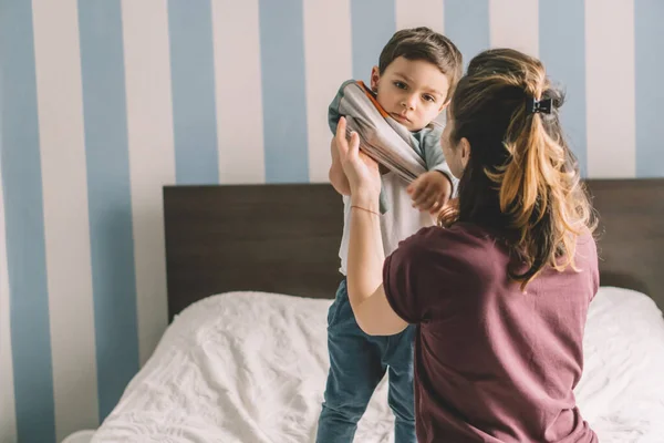 Rückansicht einer Mutter, die ihren entzückenden Sohn im Schlafzimmer anzieht — Stockfoto