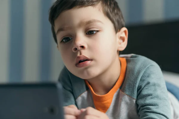 Selective focus of concentrated boy looking online lesson on smartphone in bed — Stock Photo