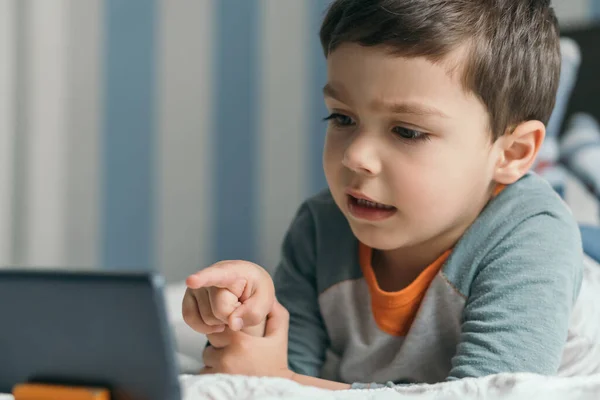 Selektiver Fokus des kleinen Jungen, der mit dem Finger auf das Smartphone zeigt, während er im Bett liegt — Stockfoto