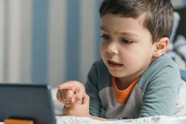 Messa a fuoco selettiva del bambino che punta con il dito lo smartphone mentre è sdraiato a letto — Foto stock