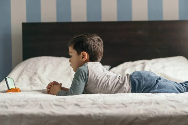 Seitenansicht des niedlichen Jungen, der auf dem Bett liegt und Online-Unterricht auf dem Smartphone anschaut — Stockfoto