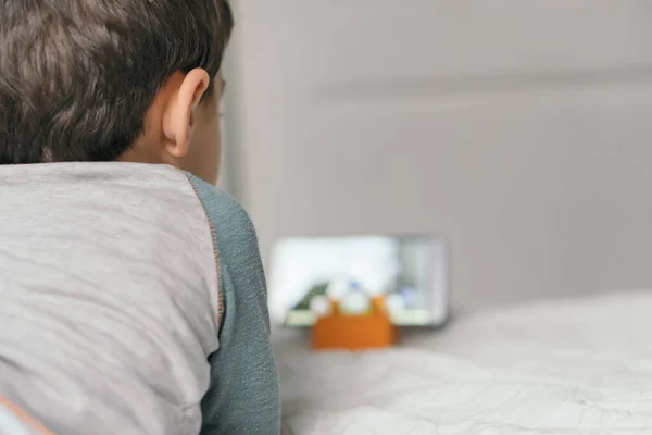 Вид сзади маленького мальчика, который смотрит веб-бинар для детей на смартфоне, лежа на кровати — стоковое фото