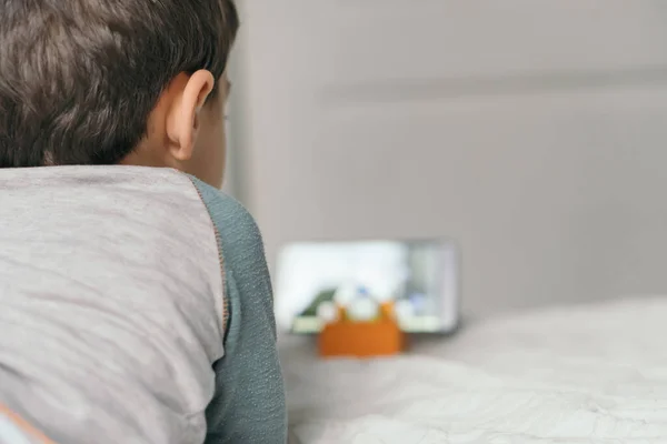 Vue arrière du petit garçon regardant webinaire pour les enfants sur smartphone tout en étant couché sur le lit — Photo de stock