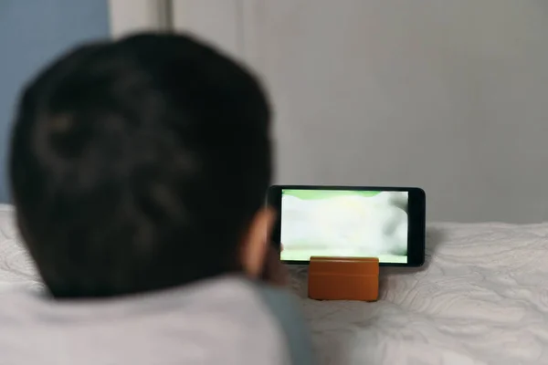 Vista posterior del niño viendo dibujos animados educativos en el teléfono inteligente mientras está acostado en la cama - foto de stock