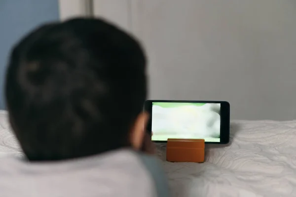 Back view of child watching educational cartoon on smartphone while lying on bed — Stock Photo