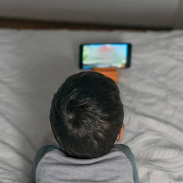 Вид сверху на мальчика, смотрящего обучающий мультфильм на смартфоне, лежа на кровати — стоковое фото