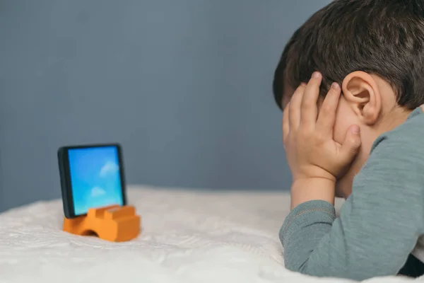 Kleiner Junge berührt Kopf, während er auf dem Smartphone Zeichentrickfilm anschaut, während er auf dem Bett liegt — Stockfoto