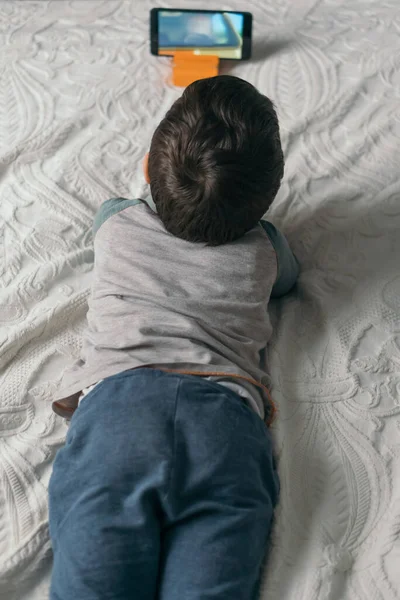 Vista superior de niño pequeño viendo webinar en el teléfono inteligente mientras está acostado en la cama - foto de stock