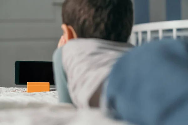 Вид сзади мальчика, смотрящего на смартфон с затемненным экраном, лежа на кровати — стоковое фото