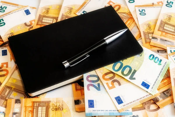 Siyah kareli bir defterin üzerinde siyah tekerlekli kalem. Arka planda Mani Euro banknotu var. İş konsepti.