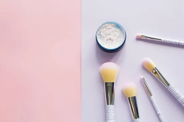 Five makeup brushes with lettering on the handle and mineral powder in a blue jar on pink and purple background. Have copy space.