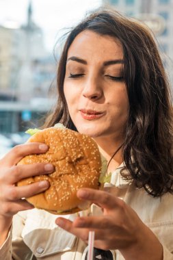 Şık giyinmiş güzel, genç bir kız kafede otururken büyük bir fast food hamburgerinin tadını çıkarıyor..