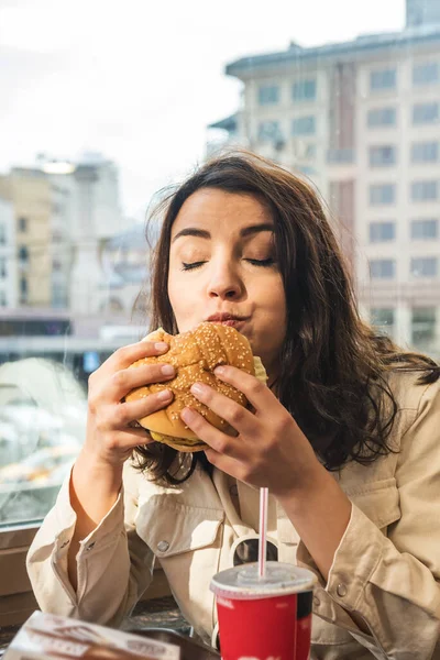 Beautiful young cute girl in fashionable clothes enjoys a big  fast food hamburger and drink while sitting in a cafe.