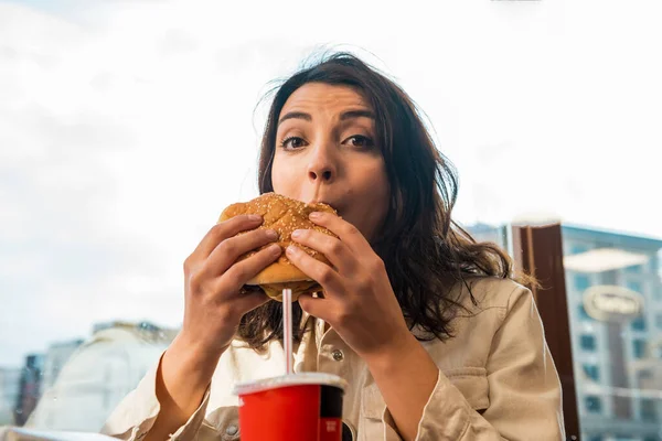 ファッショナブルな服を着た美しい若いかわいい女の子は カフェに座っている間に大きなファーストフードのハンバーガーと飲み物を楽しんでいます — ストック写真