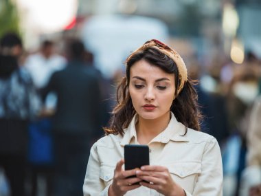 Moda kıyafetleri içindeki güzel genç kız kalabalık caddelerde yürürken akıllı telefon kullanıyor. Kalabalığın arasında bir kız telefonunu kullanıyor..