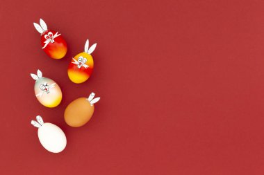 Beyaz, kahverengi ve renkli paskalya yumurtaları. Komik tavşan kulakları ve kırmızı arka planda çizgi film yüzleri var.