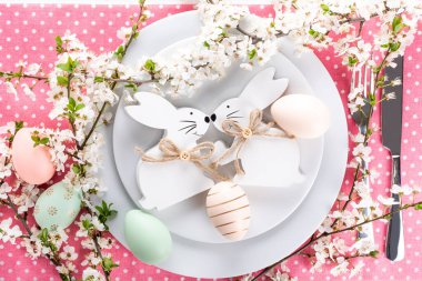 Paskalya sofrası, tatil masası dekorasyon fikri, beyaz tabaklar, çatal bıçak takımı, renkli pastel boya Paskalya yumurtaları, şenlikli dekor ve taze bahar çiçekleri ile düz konum.