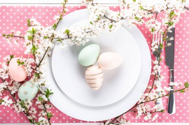 Paskalya sofrası, tatil masası dekorasyon fikri, beyaz tabaklar, çatal bıçak takımı, renkli pastel boya Paskalya yumurtaları, şenlikli dekor ve taze bahar çiçekleri ile düz konum.