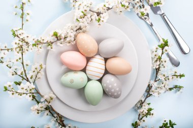Paskalya sofrası, tatil masası dekorasyon fikri, beyaz tabaklar, çatal bıçak takımı, renkli pastel boya Paskalya yumurtaları, şenlikli dekor ve taze bahar çiçekleri ile düz konum.