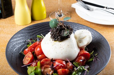 Çeri domates salatası ve roka roka yaprağı üzerine Burrata peyniri kesesi, üstünde zeytin ezmesi, siyah tabak ve restoran masası seti, çatal bıçak arka planı. Mozzarella geleneksel İtalyancası