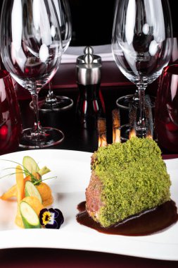 Bir parça kuzu pirzolası, yeşil otla kızartılmış, ekmek, peynir kırıntıları, tatlı patates püresi ve bebek sebzeleriyle servis edilmiş, kırmızı şarap sosu, şık bir restoranda, beyaz tabakta.