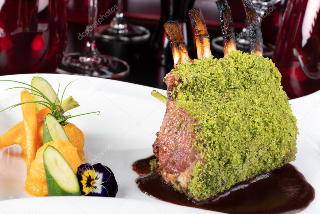Piece of lamb rack with bone roasted with green herb, bread, cheese crumbs cover, served with sweet potatoes puree and baby vegetables, red wine sauce, in a restaurant elegant setting, on white plate