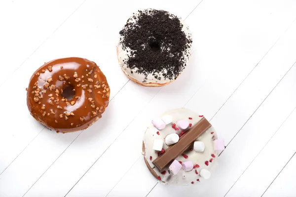 Three melted glazed with dark and white chocolate, marshmallows, biscuit crumbles, caramel sugar coated donuts on white wood board