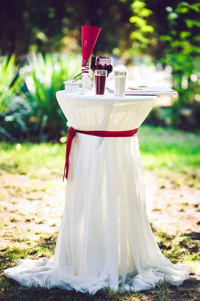 Uma mesa na cerimônia de casamento, decorada com um pano branco com um arco vermelho. Na mesa estão copos de recém-casados, taças de areia e muito mais. . — Fotografia de Stock