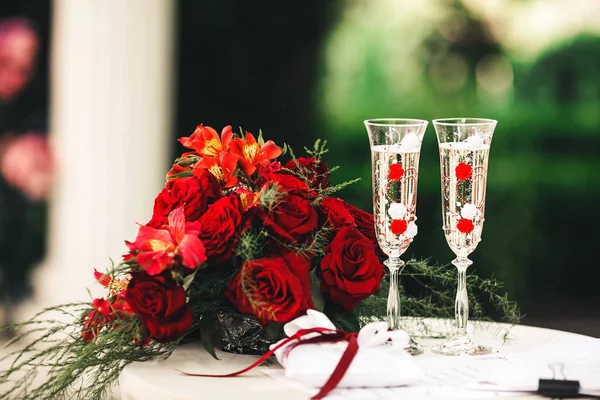 Um buquê vermelho, belos copos com champanhe e um travesseiro para anéis de casamento estão sobre a mesa — Fotografia de Stock