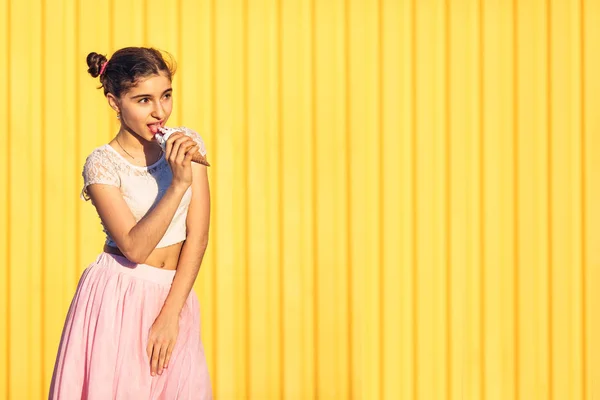 Chica joven en falda rosa está comiendo helado sobre fondo amarillo de la pared . — Foto de Stock