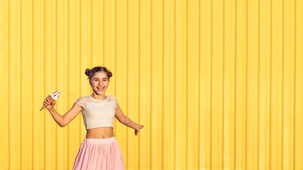 Feliz chica con estilo en falda rosa con helado en la mano y sonriendo contra el fondo amarillo de la pared . — Foto de Stock