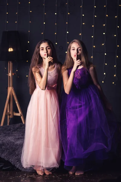 Veselé přítelkyně. Dvě dívky v růžové a fialové šaty ukazují, studio na tmavém pozadí. Kopírovat prostor. — Stock fotografie