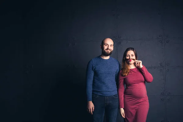 Homem barbudo sorri, e mulher grávida faz bigode fora do cabelo no estúdio em fundo escuro. Espaço de cópia . — Fotografia de Stock
