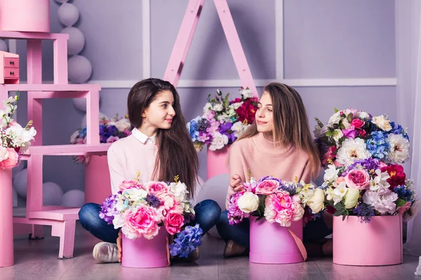 Dvě krásné dívky v džíny a růžový svetr ve studiu s dekorem květin v koších. — Stock fotografie