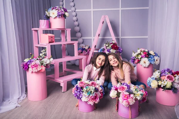 Dvě krásné dívky v džíny a růžový svetr ve studiu s dekorem květin v koších. — Stock fotografie
