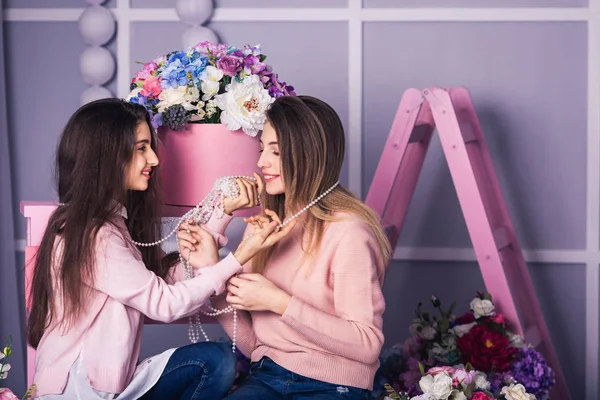 Dvě krásné dívky v džíny a růžové svetry se drží korálky ve studiu s dekorem květin v koších. — Stock fotografie
