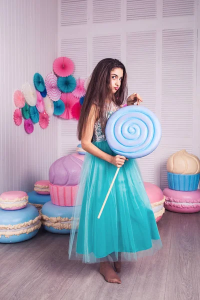 Mooi meisje in blauwe jurk Houd in de hand enorme snoep in studio met decor van bitterkoekjes. Kopiëren van ruimte. — Stockfoto