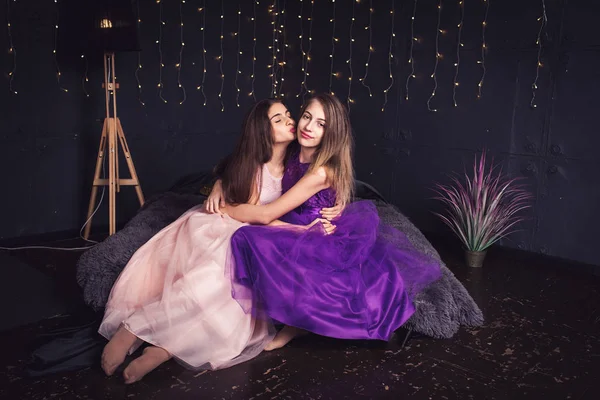 Veselá přítelkyně. Dvě dlouhé vlasy dívky v růžové a fialové šaty v studio na tmavém pozadí s rozostření pozadí. Kopírovat prostor. — Stock fotografie