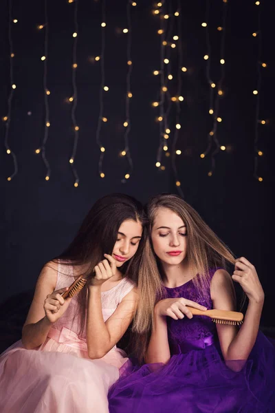 Veselá přítelkyně. Dvě dívky hřeben vzájemně si vlasy, sedí vedle sebe v studio na tmavém pozadí. Kopírovat prostor — Stock fotografie