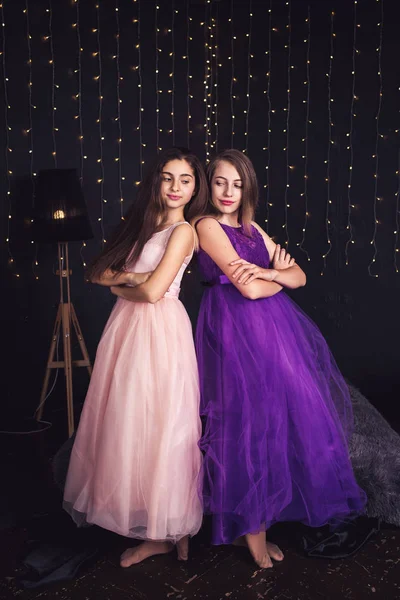 Veselá přítelkyně. Dvě dlouhé vlasy dívky v růžové a fialové šaty v studio na tmavém pozadí s rozostření pozadí. Kopírovat prostor. — Stock fotografie