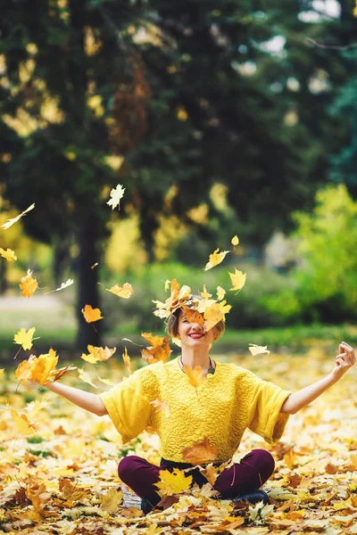 Girl in autumn sits on grass and cheerfully casts up yellow leaves. Life style. — Stock Photo, Image
