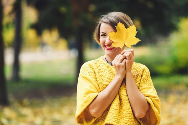 Menina sorridente em suéter amarelo no parque no outono fecha os olhos com folha de bordo . — Fotografia de Stock