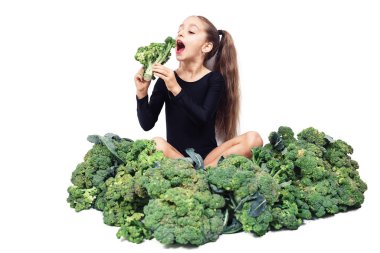 little girl gymnast in leotard sits among broccoli and eats it. Baby Vegan Isolated On White Background. clipart