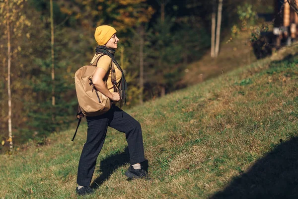 Young tourist girl with backpack rises in autumn on hill in forest in mountains. — Stock Photo, Image