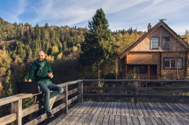 Sakallı adam tırabzanda oturur ve dizüstü bilgisayarda çalışır. Dağlarda tatilde olan serbest yazar hipster..